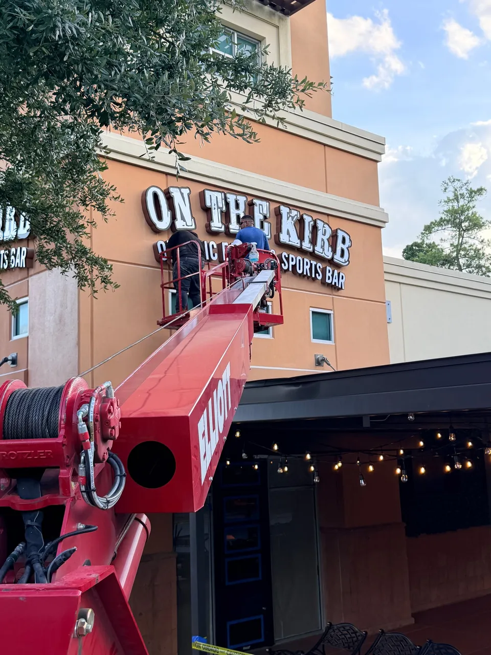 Two installers in the bucket of a red Elliott boom truck mounting white channel letters reading ON THE KIRB above a patio at a two-story orange stucco restaurant
