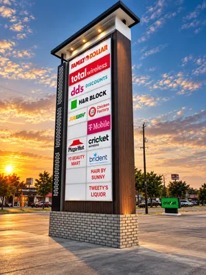 Tall multi-tenant pylon sign at sunset with a lit cabinet of fourteen tenant panels in two columns, dark wood-look cladding, a brick base and downlights under the cap, in a retail center parking lot